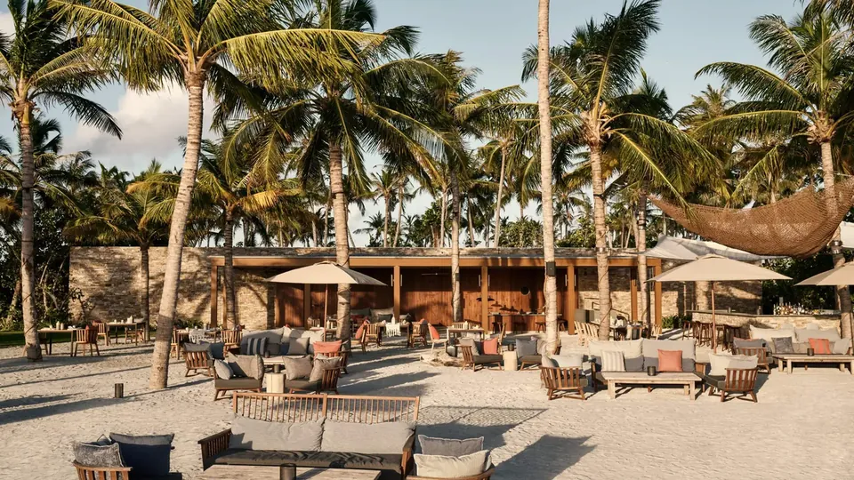 A beach restaurant with wooden tables and chairs under palm trees on a sunny day