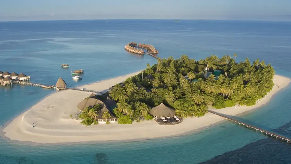 Una pequeña isla con palmeras y playa de arena blanca, con un embarcadero y villas sobre una laguna turquesa