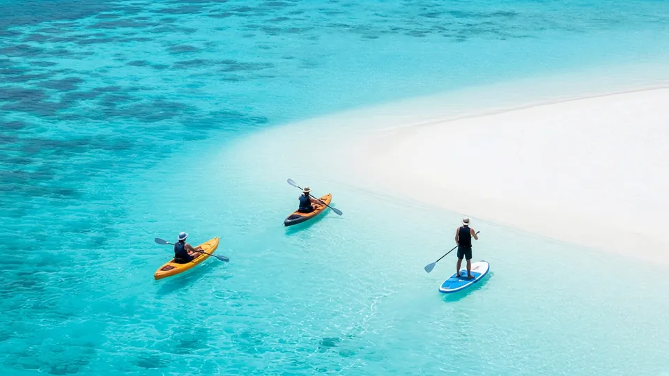 Deux kayakistes et une personne en paddle, pagaie en main, dans un lagon turquoise limpide à côté d’un banc de sable blanc, vus d’en haut