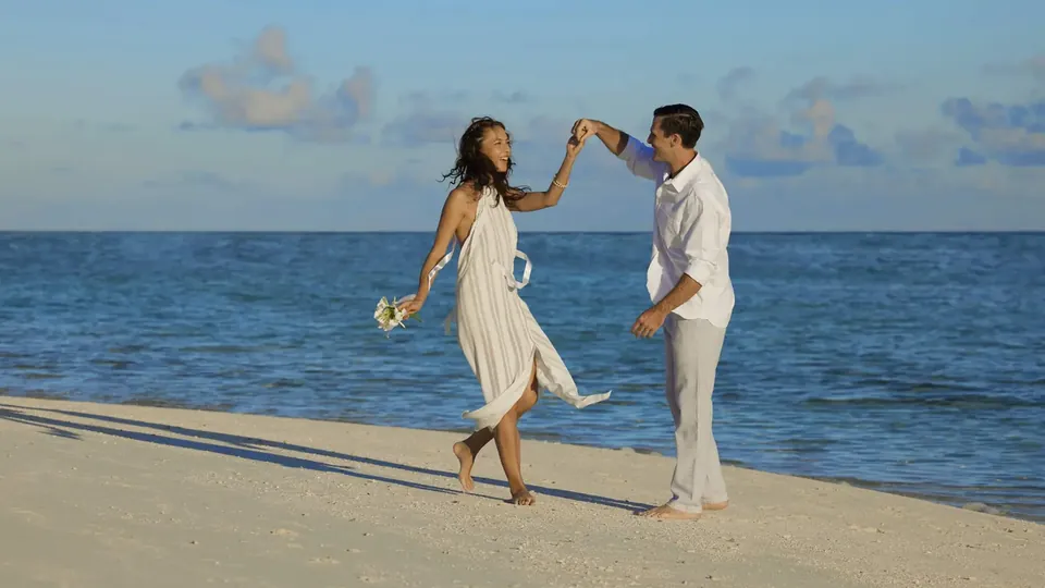 Um casal com roupas brancas de verão dançando descalço em uma praia ensolarada