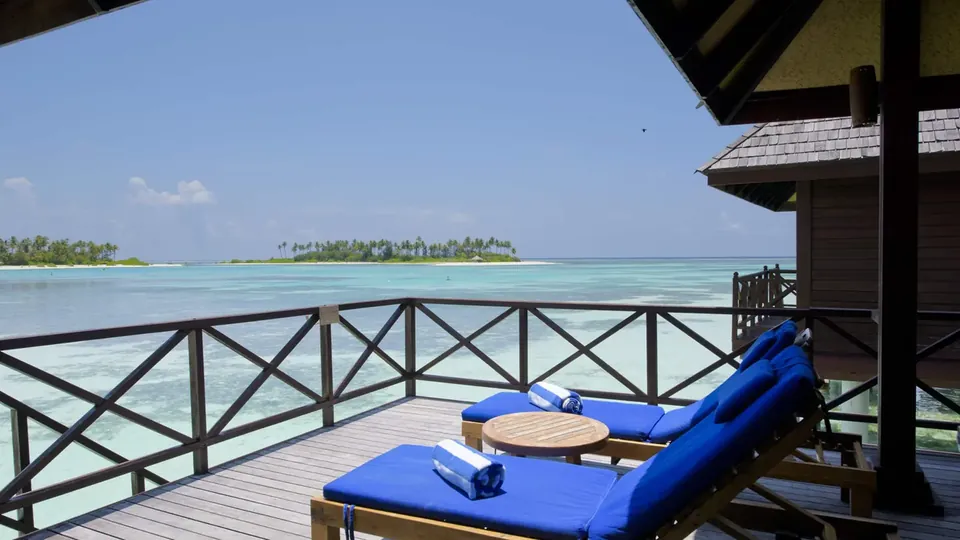 A sunny overwater villa deck with blue sun loungers facing the lagoon
