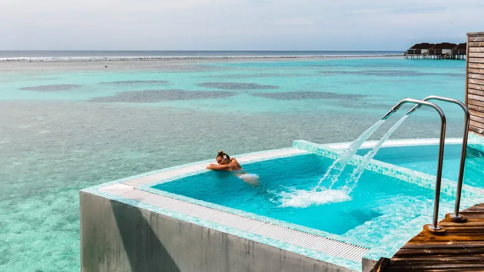 A water villa deck with a jetted tub and loungers above a calm blue lagoon